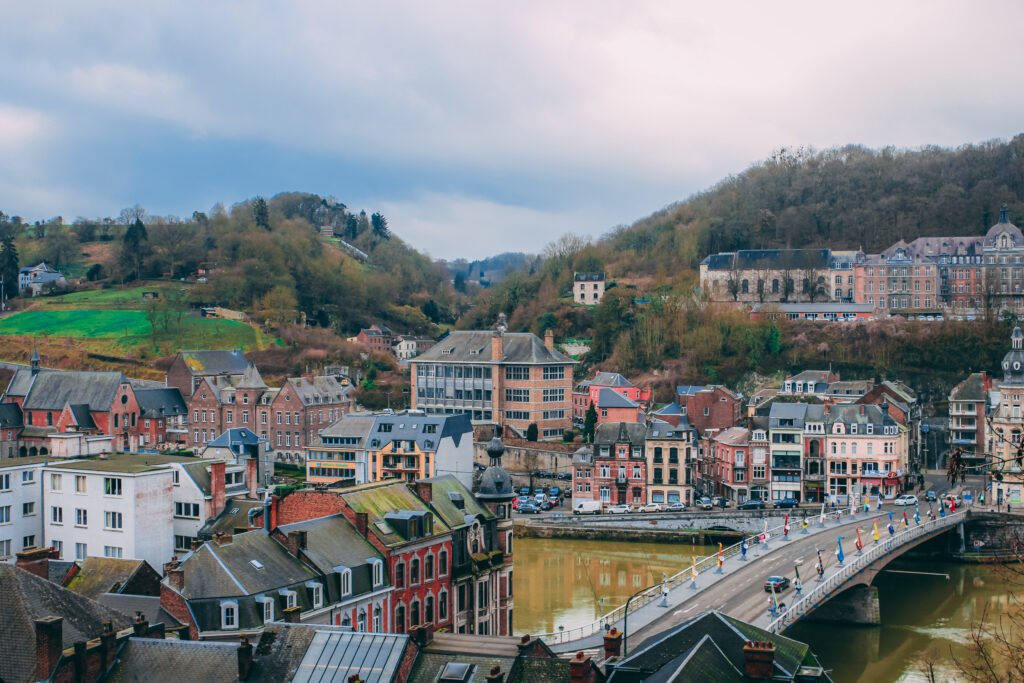 Vista aérea de Dinant, Bélgica, com edifícios históricos coloridos, uma ponte sobre o rio Meuse e colinas arborizadas ao fundo.