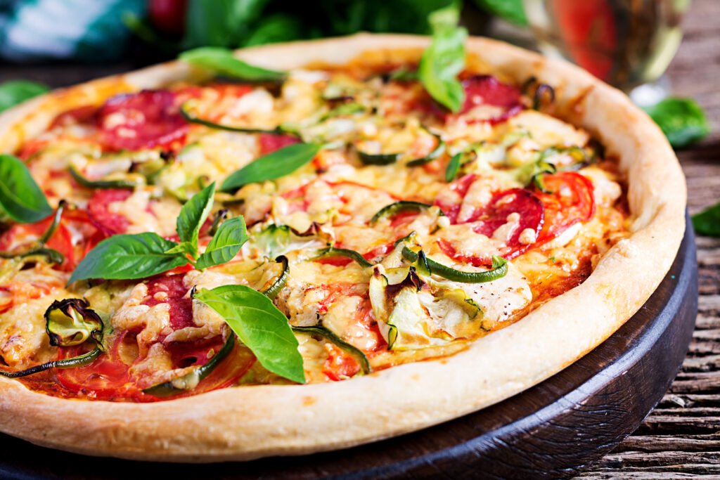 Delicious Italian pizza topped with chicken, salami, zucchini, tomatoes, and fresh herbs, served on a rustic wooden table — traditional Italian cuisine at its finest.