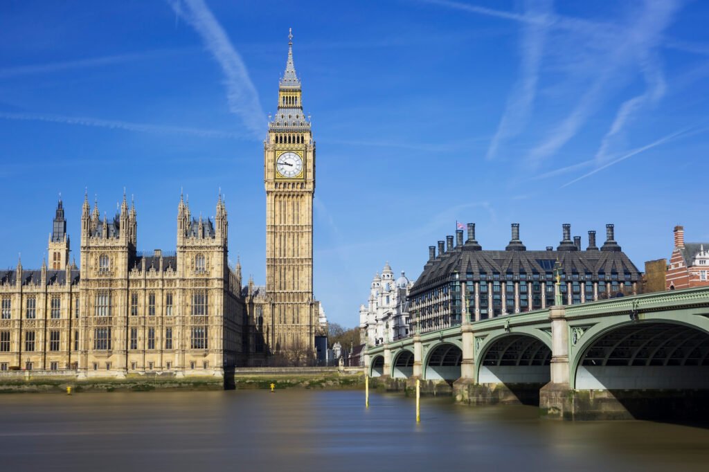 View of Big Ben in the background on a clear blue sky day, with a calm river in the foreground and a bridge on the right, capturing the sunny urban landscape of London, one of the most beautiful cities in England.