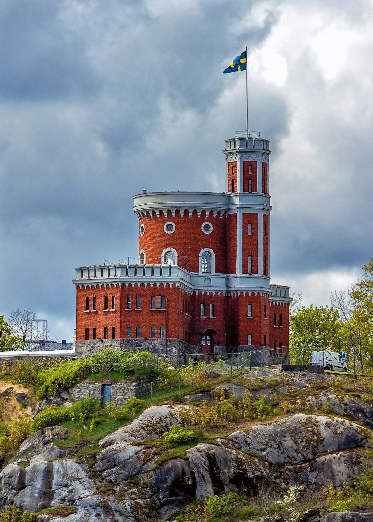 Scenic view of a red castle with white details in one of the most beautiful cities in Sweden, Stockholm, surrounded by lush greenery, rocks, and a partly cloudy blue sky, featuring a white van beside the castle and trees in the background.