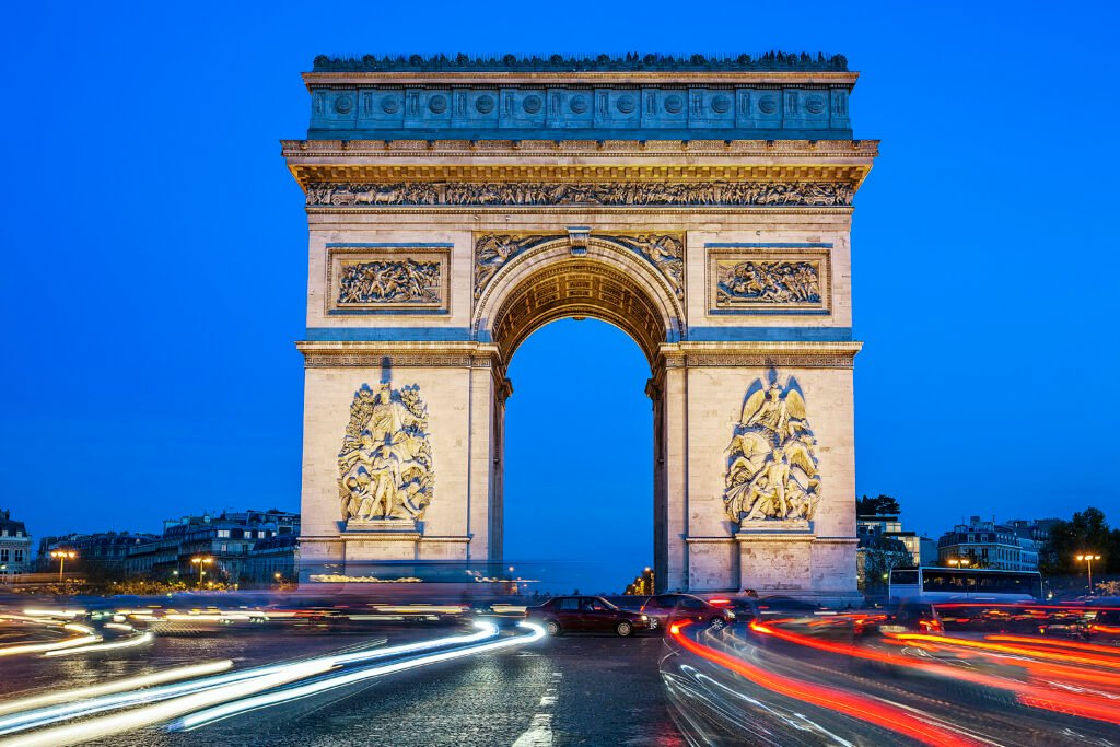 Arc de Triomphe in Paris at night, beautifully illuminated with light trails from cars circling the busy Champs-Élysées intersection.