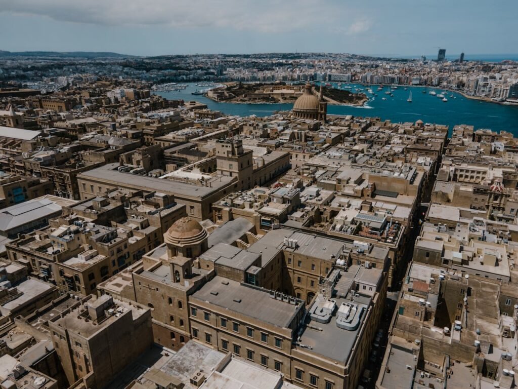 Aerial image of Il-Belt Valletta, the historic capital of Malta, with its golden stone buildings and narrow streets. In the background, a body of water that looks like an urban river or inlet, contrasting with the blue of the Mediterranean Sea.
