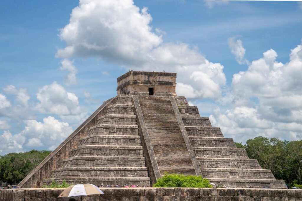 Aerial view of the Kukulcán pyramid with staircases at Chichén Itzá, Yucatán, Mexico, under a blue sky with white clouds and surrounded by green vegetation. Chichén Itzá is one of the most famous attractions in one of the most beautiful Latin American countries and a UNESCO World Heritage site.
