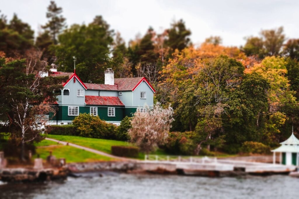 Charming green and white house with a red roof by the lake, surrounded by trees and lush vegetation, featuring a pier that ends in a small white cylindrical castle-shaped building.