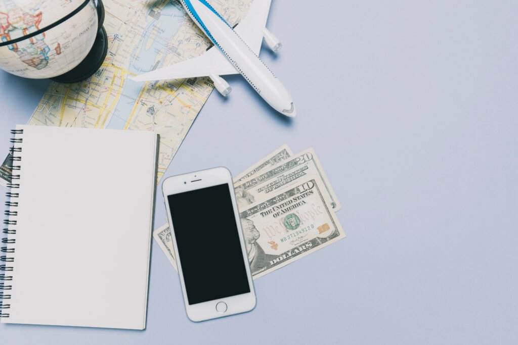 Table with travel items: a planner, a small decorative globe, a map, a smartphone (possibly an iPhone), three U.S. dollar bills (a $20, a $10, and one partially visible), and a miniature airplane.