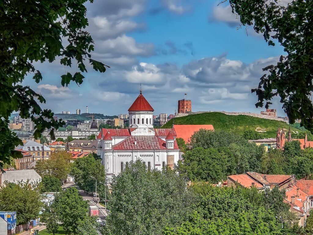 Aerial view framed between two trees, featuring a central building with castle-like towers — possibly a church — surrounded by smaller buildings and lush greenery, in a typical Lithuanian landscape.