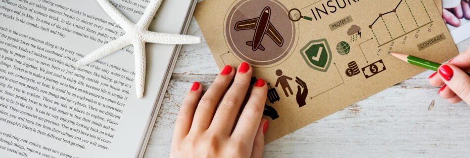 Hands with red nail polish holding a travel insurance brochure with icons and charts, placed on a white wooden surface next to an open book, a starfish, and a camera.