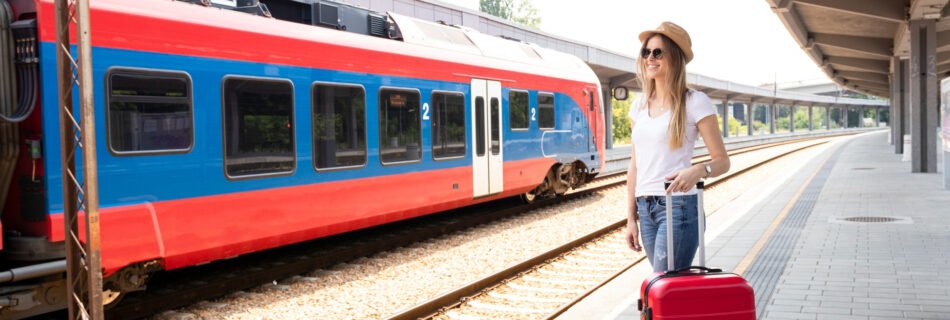 Young woman standing on a sunny train platform in Europe, wearing blue jeans, a white t-shirt, white sneakers, and a brown hat, smiling next to a red and blue train with a red suitcase.