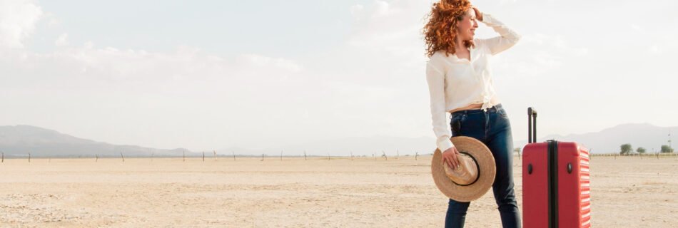 Woman traveling alone with curly red hair, standing in a desert landscape with a red suitcase and a straw hat in hand, looking into the distance.