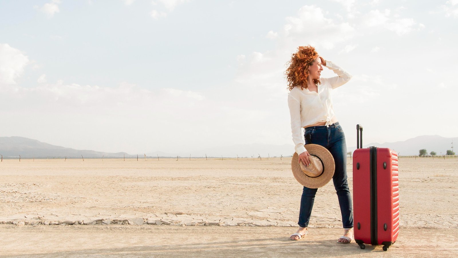 Woman traveling alone with curly red hair, standing in a desert landscape with a red suitcase and a straw hat in hand, looking into the distance.