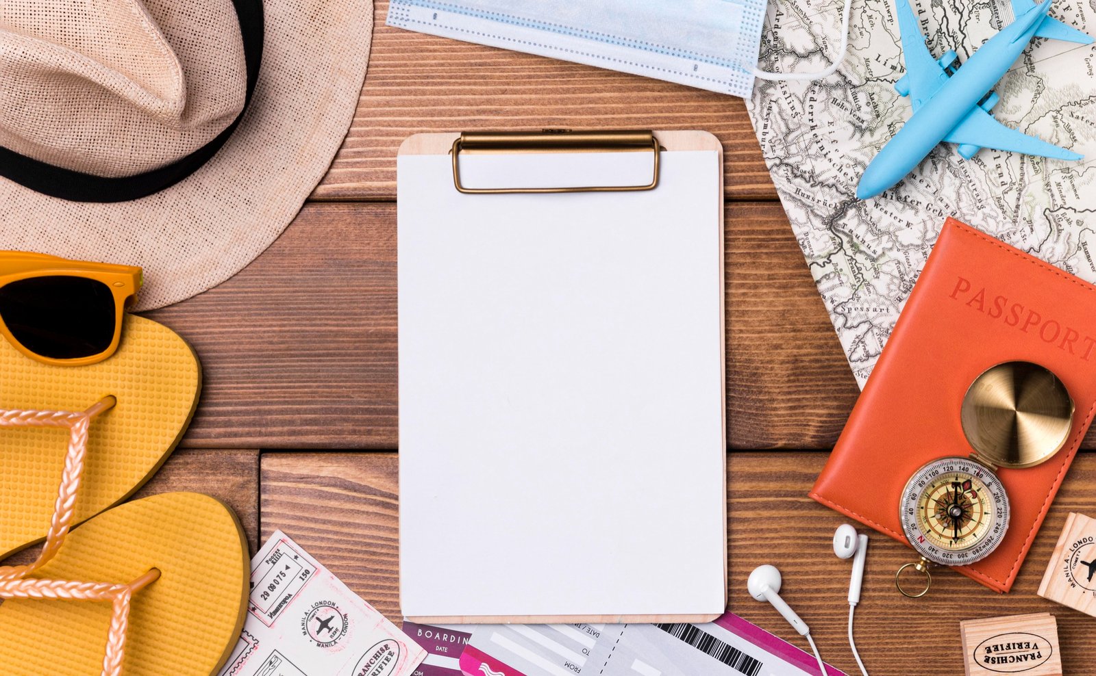 Flat lay of travel essentials on a wooden surface including a straw hat, sunglasses, flip-flops, a blank clipboard, boarding passes, a face mask, a passport holder with a compass, and a toy airplane on a vintage map.