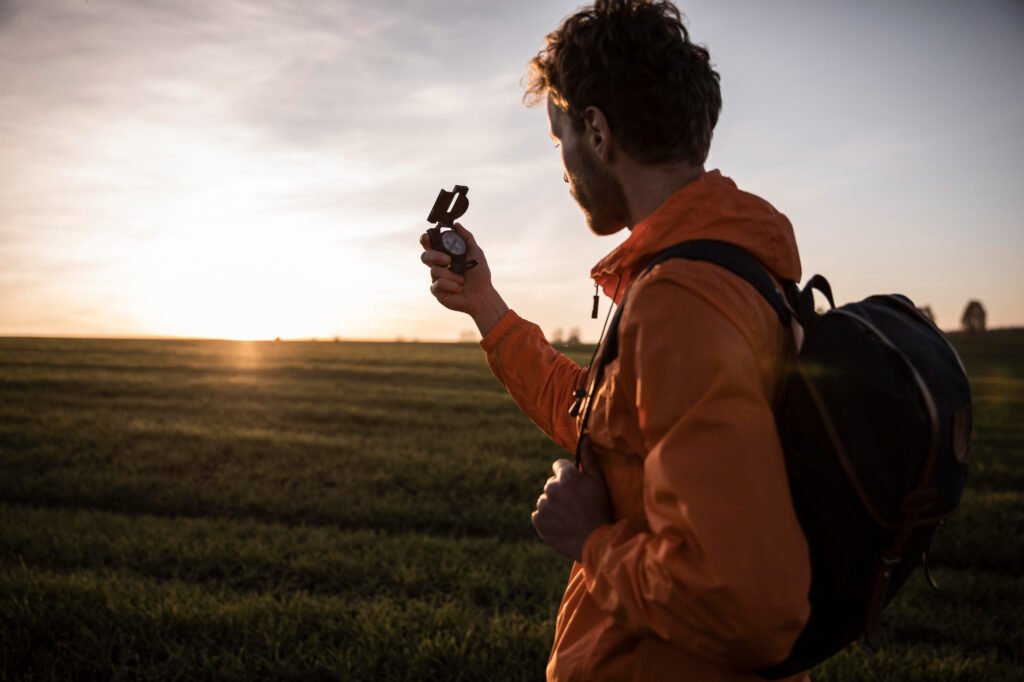 Man traveling alone wearing an orange jacket and backpack, holding a compass while standing in a grassy field at sunset.