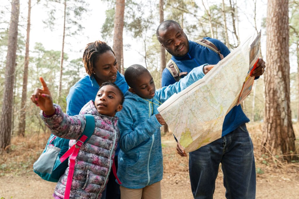 A family of four, wearing casual hiking clothes and backpacks, gathers in a forest as they examine a large paper map together. The father holds the map while looking at it, the mother and two children—one of whom is also pointing—look engaged and curious. The tall trees and natural light suggest a peaceful outdoor adventure.