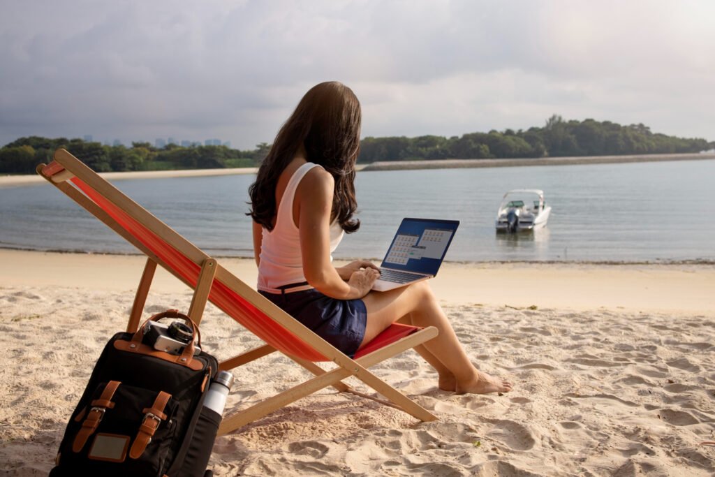 Woman sitting alone on a beach chair with a laptop on her lap, working remotely near the ocean. A black backpack and water bottle sit beside her in the sand, while a small boat floats in the water and a tropical forest lines the horizon.