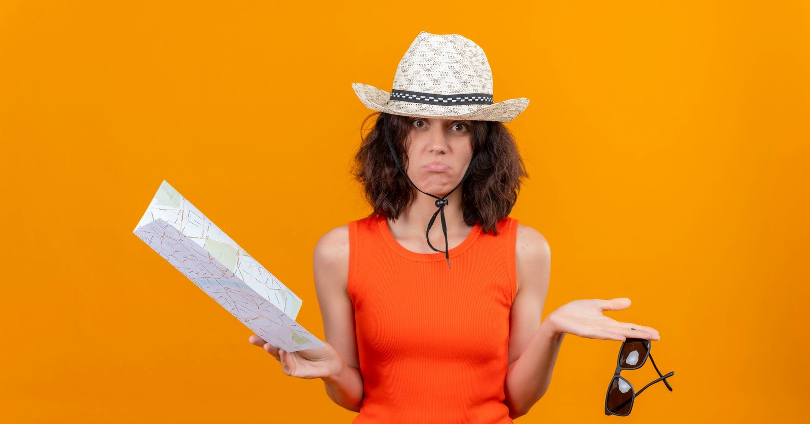 Frustrated female traveler in an orange dress and straw hat holding a map in one hand and sunglasses in the other, looking confused against an orange background — representing travel mistakes or getting lost on a trip.