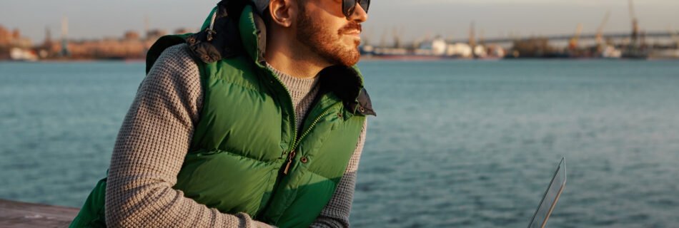 Man leaning on a wooden railing by the water, wearing sunglasses, a gray cap, and a green puffer vest, with a laptop open in front of him. The industrial harbor and blue sky in the background suggest a quiet moment during remote work travel.