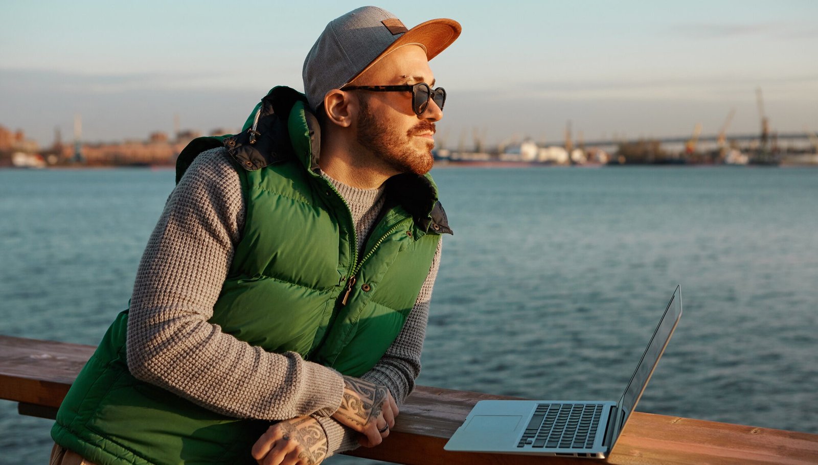 Man leaning on a wooden railing by the water, wearing sunglasses, a gray cap, and a green puffer vest, with a laptop open in front of him. The industrial harbor and blue sky in the background suggest a quiet moment during remote work travel.