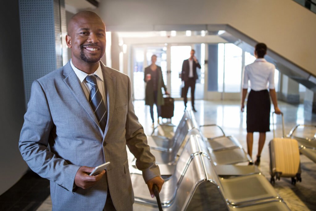 Business traveler in a gray suit smiling while standing in a modern airport terminal, holding a phone and a suitcase handle, with other passengers in the background.