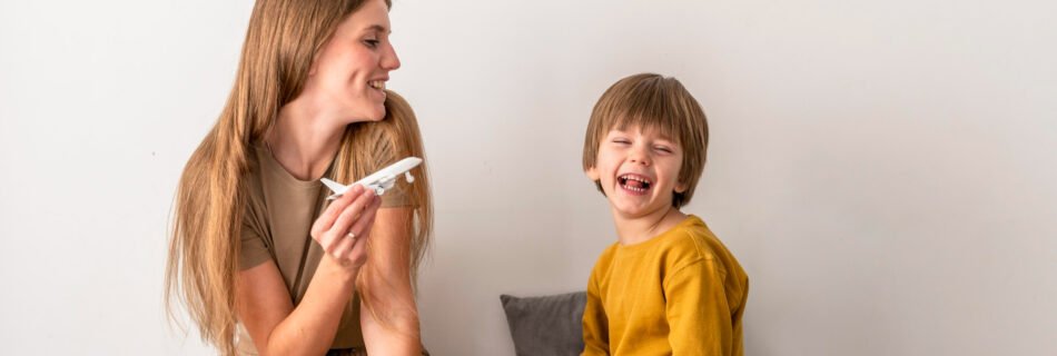 A smiling mother and her young son sit on a bed while packing an open suitcase filled with neatly folded clothes. The mother, holding a toy airplane, engages the boy in conversation, sparking excitement. The boy, dressed in a mustard-yellow shirt, laughs joyfully, capturing a lighthearted moment of preparing for a family trip.