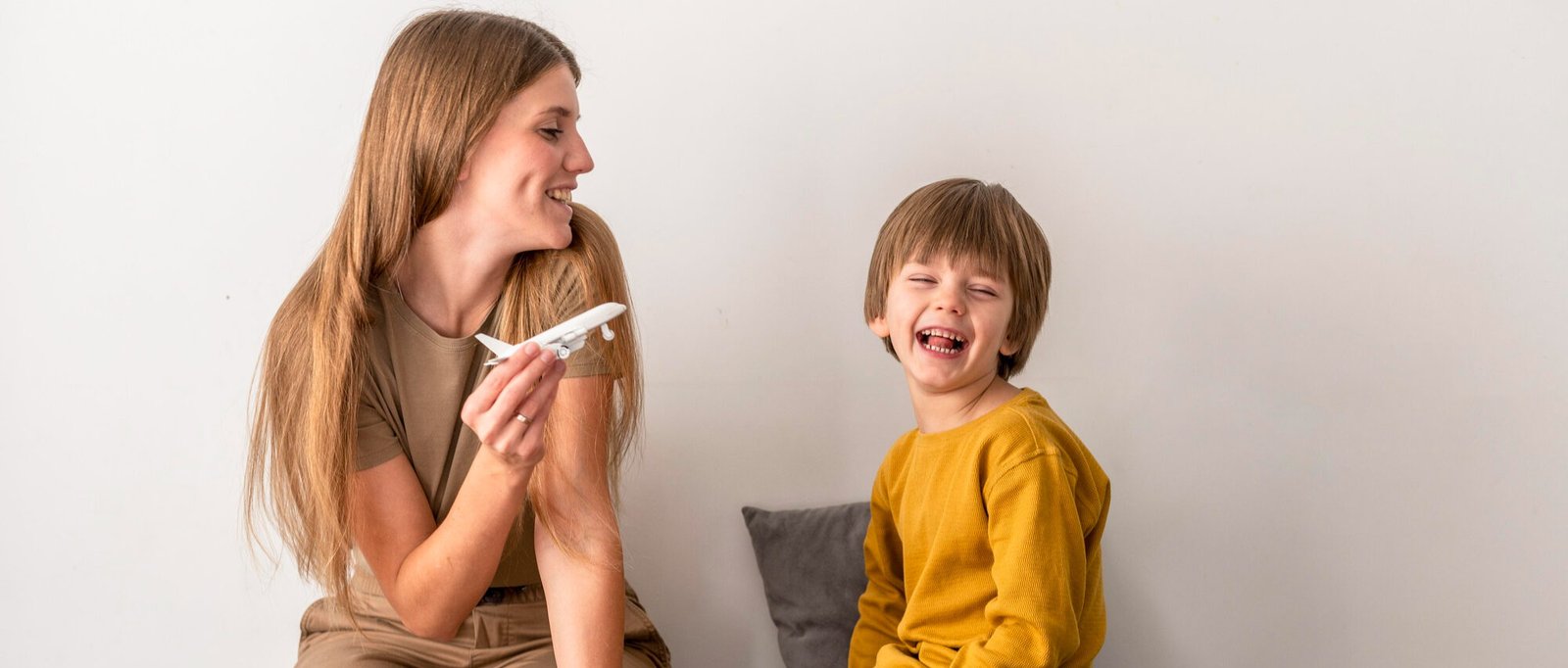 A smiling mother and her young son sit on a bed while packing an open suitcase filled with neatly folded clothes. The mother, holding a toy airplane, engages the boy in conversation, sparking excitement. The boy, dressed in a mustard-yellow shirt, laughs joyfully, capturing a lighthearted moment of preparing for a family trip.