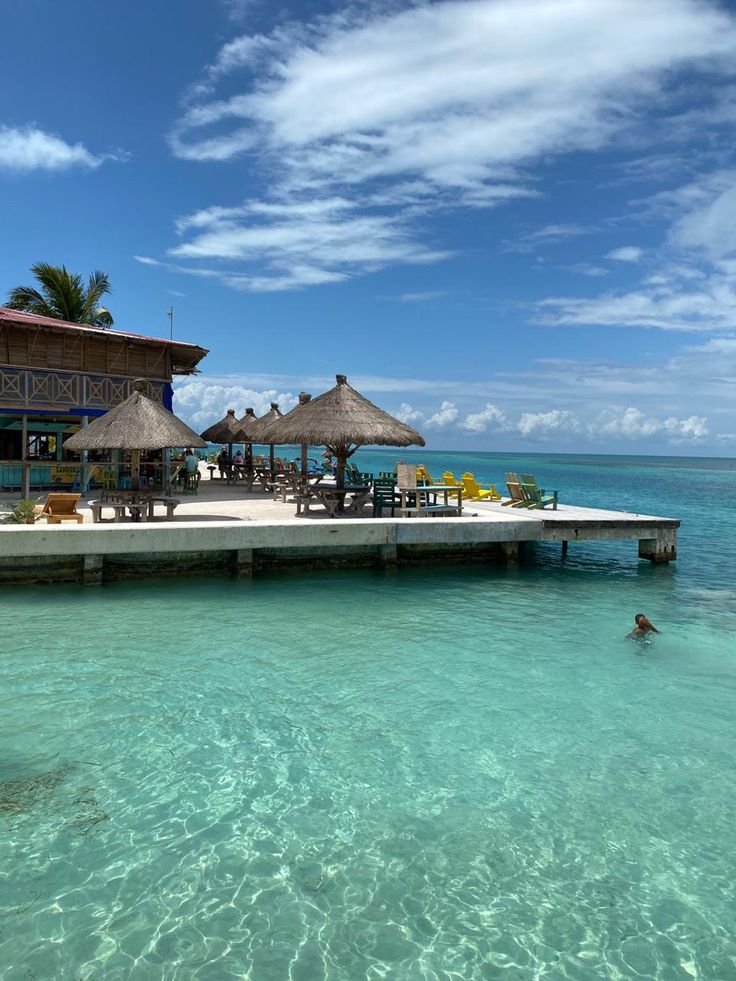 Scenic view of Caye Caulker beach in Belize with crystal-clear turquoise water, wooden pier, and palm trees under a blue sky.