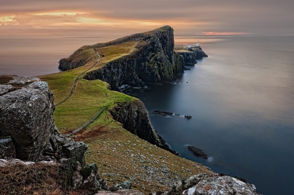 Cliffside view of Neist Point Lighthouse on the Isle of Skye, Scotland, with dramatic cliffs and ocean waves under a cloudy sky.