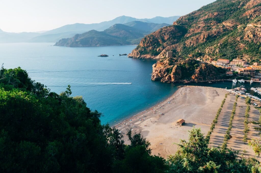 Panoramic view of Corsica, France, showing a beach with turquoise blue-green sea, mountains in the background under a cloudy sky, and a light mist on the horizon.