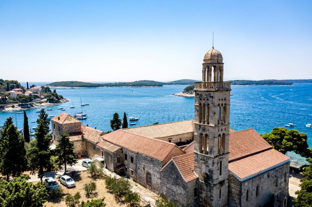 View of Hvar, Croatia, featuring an old stone house with a tower in the foreground, trees and parked cars nearby, a blue sea filled with many boats sailing, and small islands visible in the background.