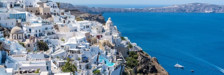 Panoramic view of Santorini, Greece, featuring iconic white houses on cliffs facing the deep blue sea under a partially cloudy sky.