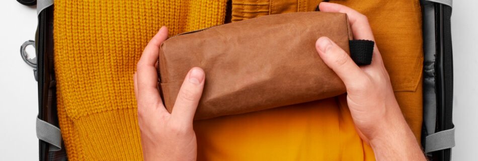 Hands placing a brown toiletry bag inside a neatly packed suitcase with yellow clothing.