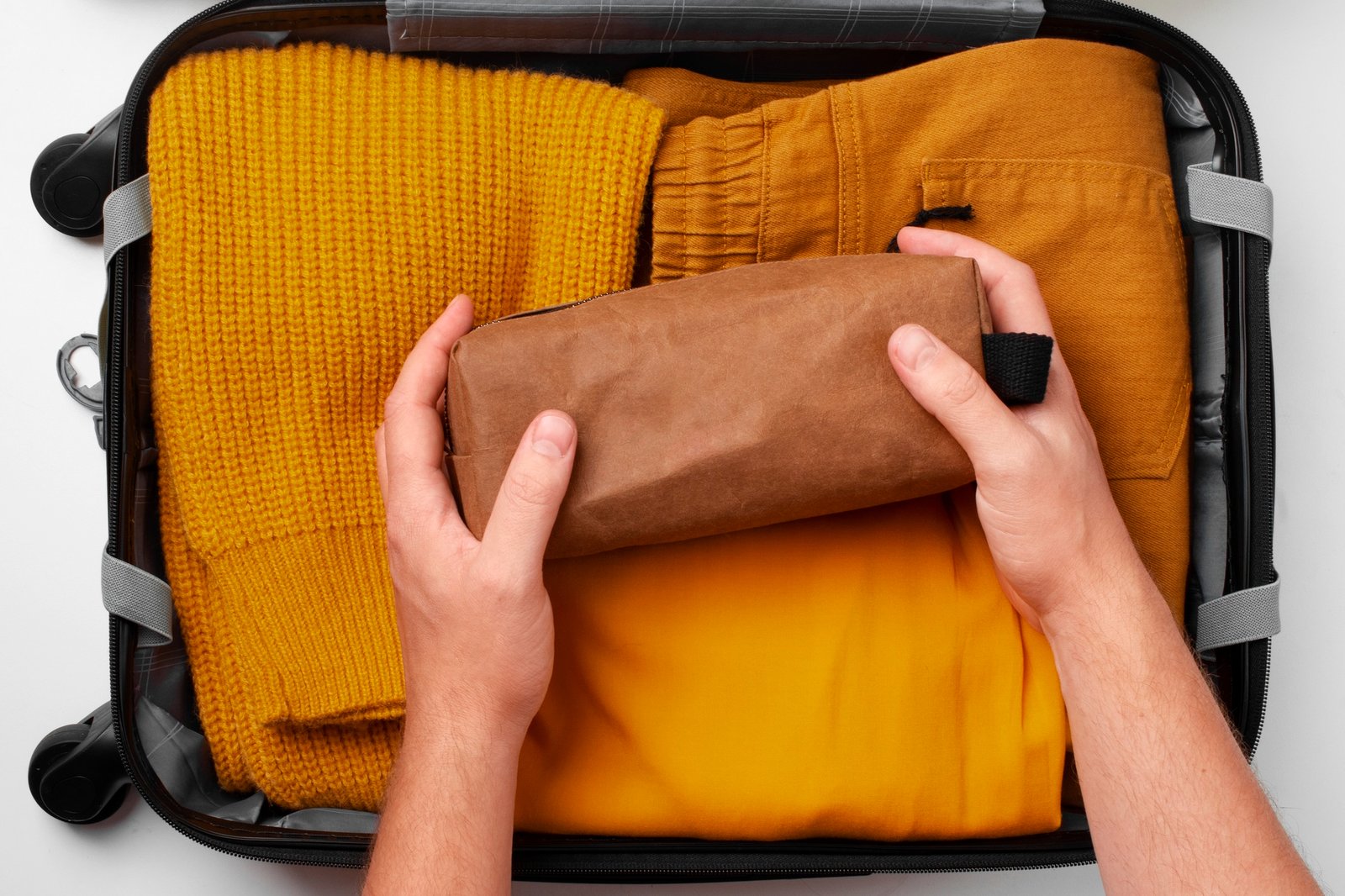 Hands placing a brown toiletry bag inside a neatly packed suitcase with yellow clothing.