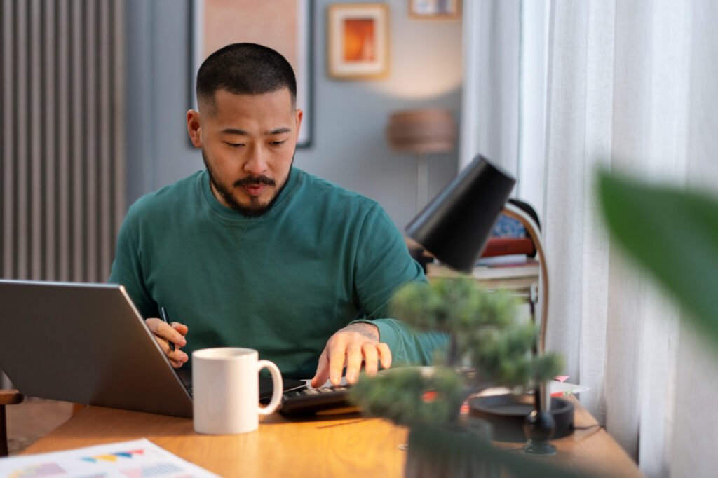 Asian man wearing a green shirt holding a pen while using a calculator, with a laptop on the table in a coworking space.