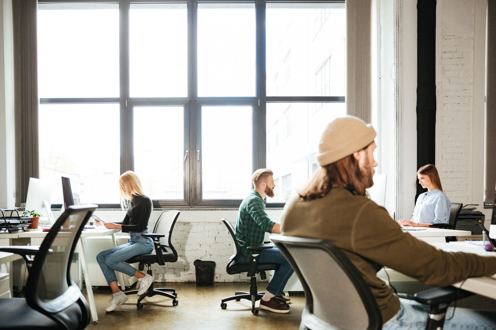 Two men and two women working in a modern coworking space, with eight large windows in the background letting in natural light and stylish, modern chairs throughout the room.