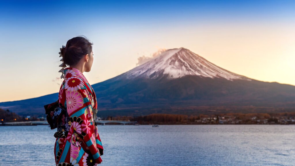 Mulher usando quimono colorido tradicional japonês, olhando para o Monte Fuji coberto de neve ao fundo durante o pôr do sol.