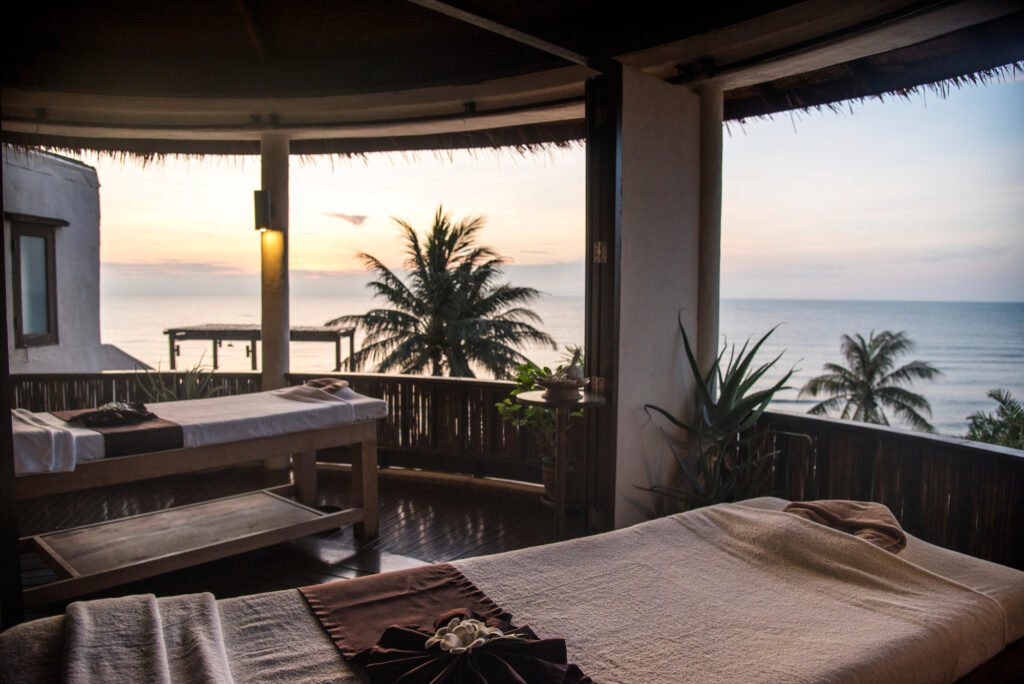 Spa room with massage tables facing the ocean, decorated with tropical plants and a stunning sunset view.