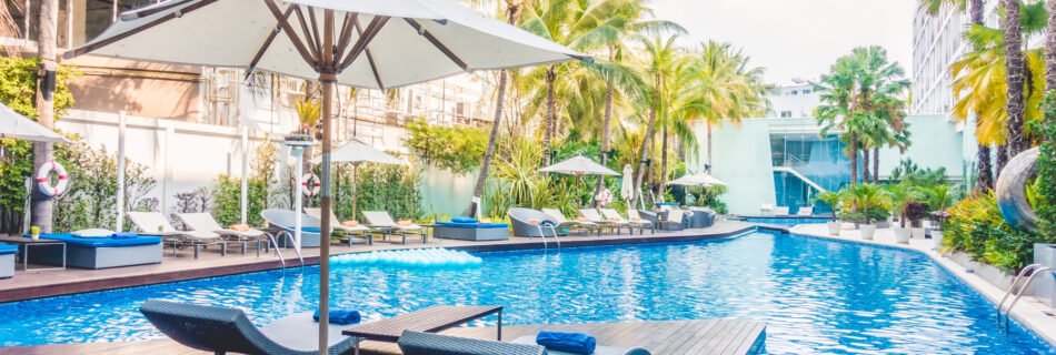 Outdoor pool area at a hotel or resort, with lounge chairs, white umbrellas, and many palm trees around on a sunny day. Perguntar ao ChatGPT