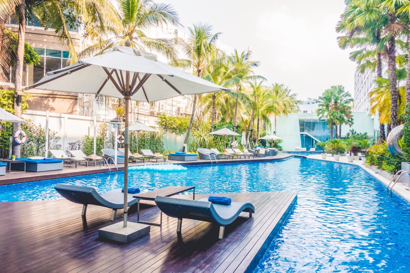 Outdoor pool area at a hotel or resort, with lounge chairs, white umbrellas, and many palm trees around on a sunny day. Perguntar ao ChatGPT
