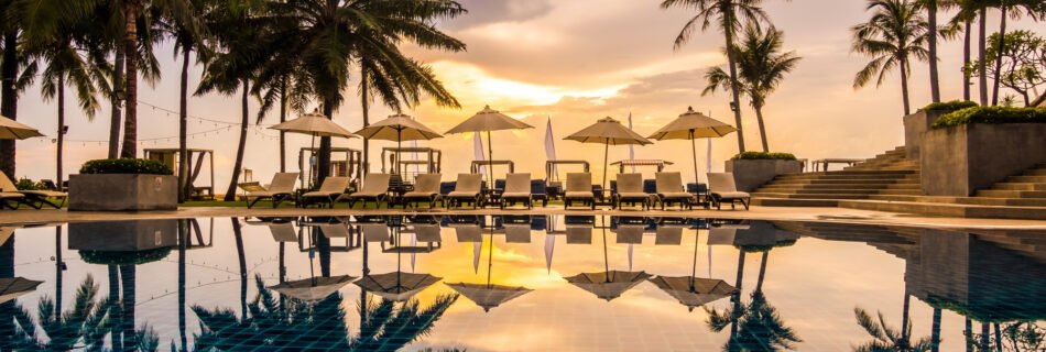 Hotel swimming pool with sun loungers and white umbrellas, surrounded by palm trees and reflecting a golden sunset on the water.