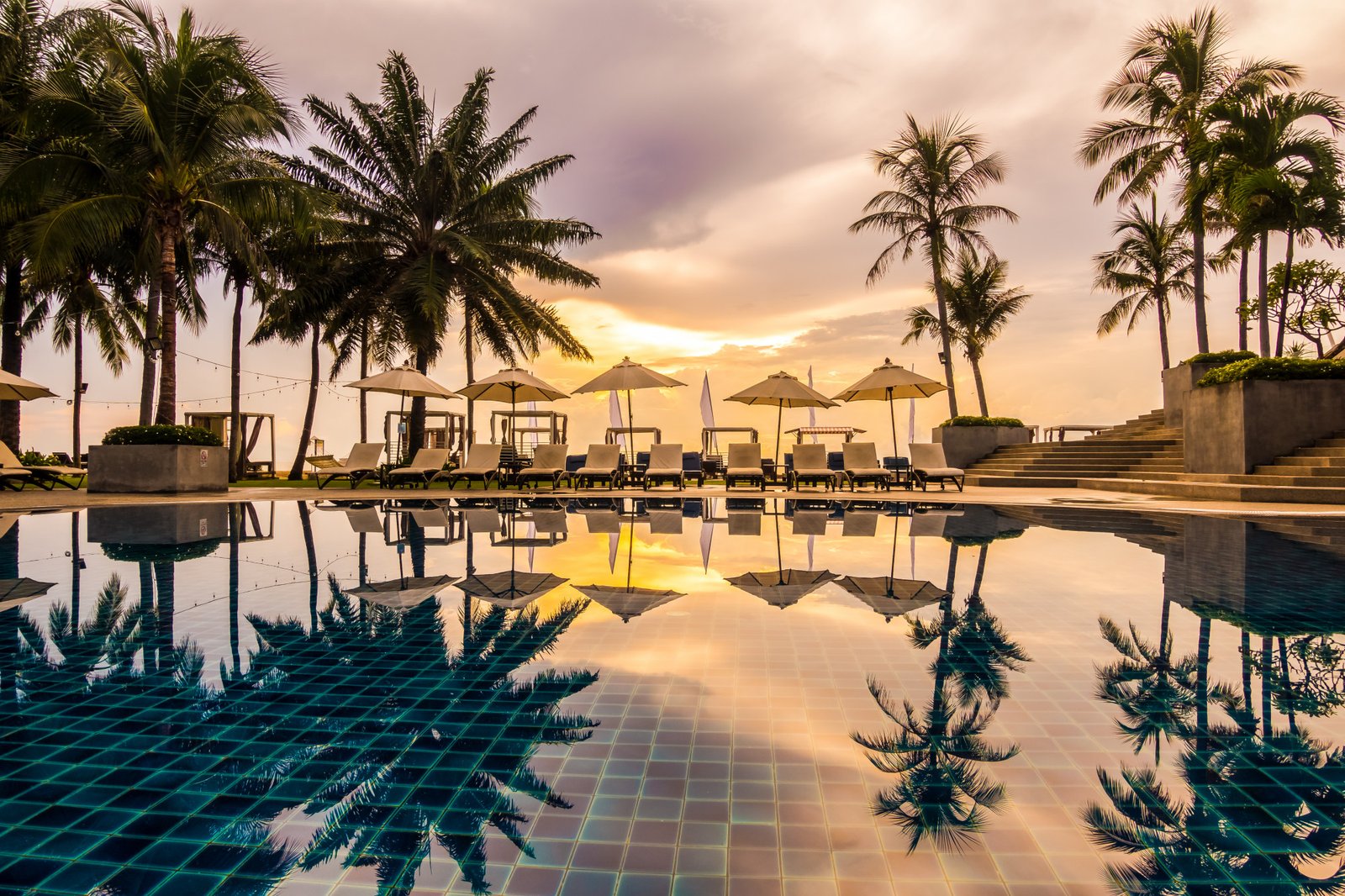 Hotel swimming pool with sun loungers and white umbrellas, surrounded by palm trees and reflecting a golden sunset on the water.