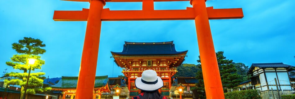 Tourist seen from behind wearing a backpack and a white hat, looking at a large red torii gate at a traditional Japanese temple at dusk.