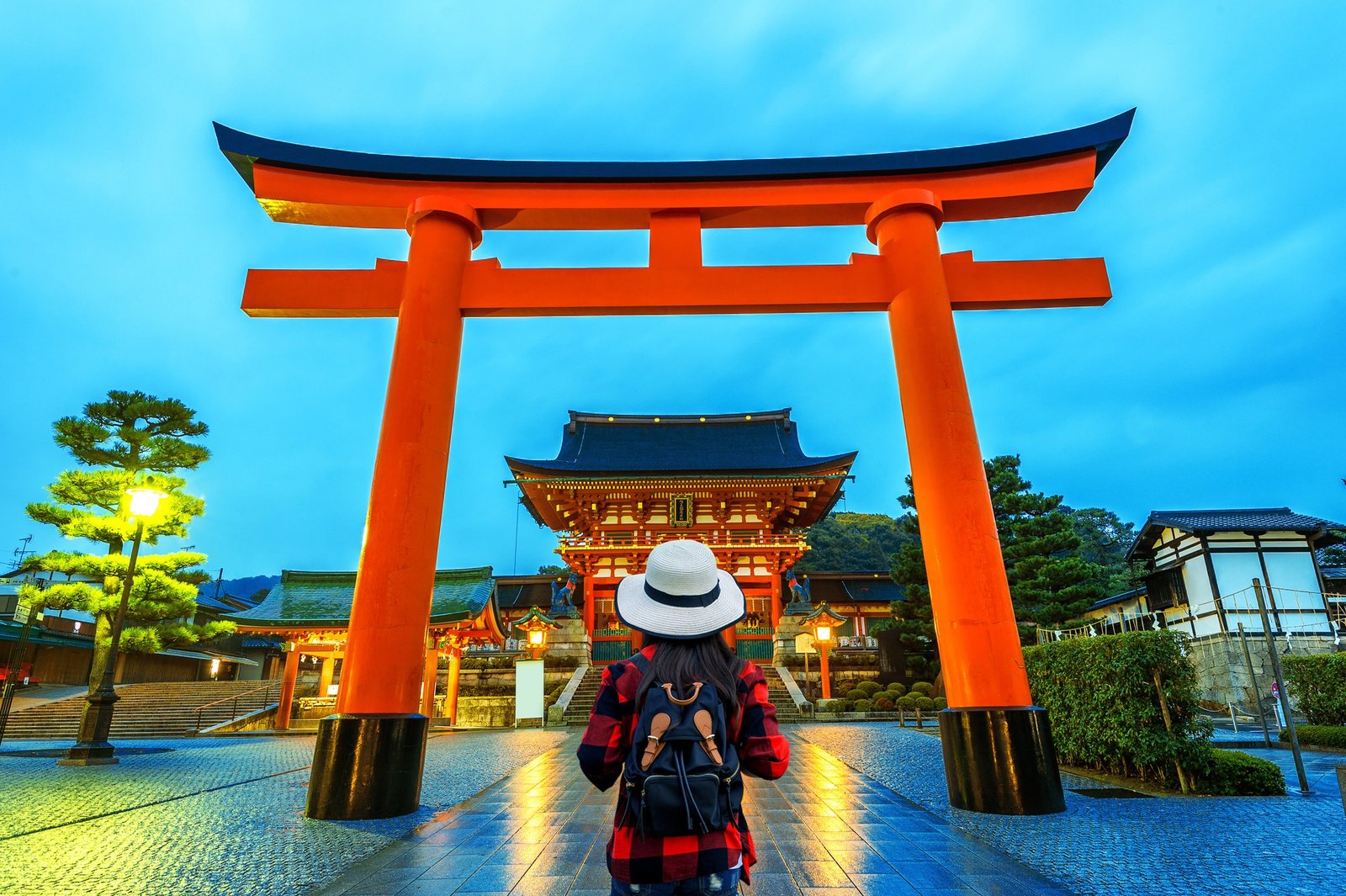 Tourist seen from behind wearing a backpack and a white hat, looking at a large red torii gate at a traditional Japanese temple at dusk.