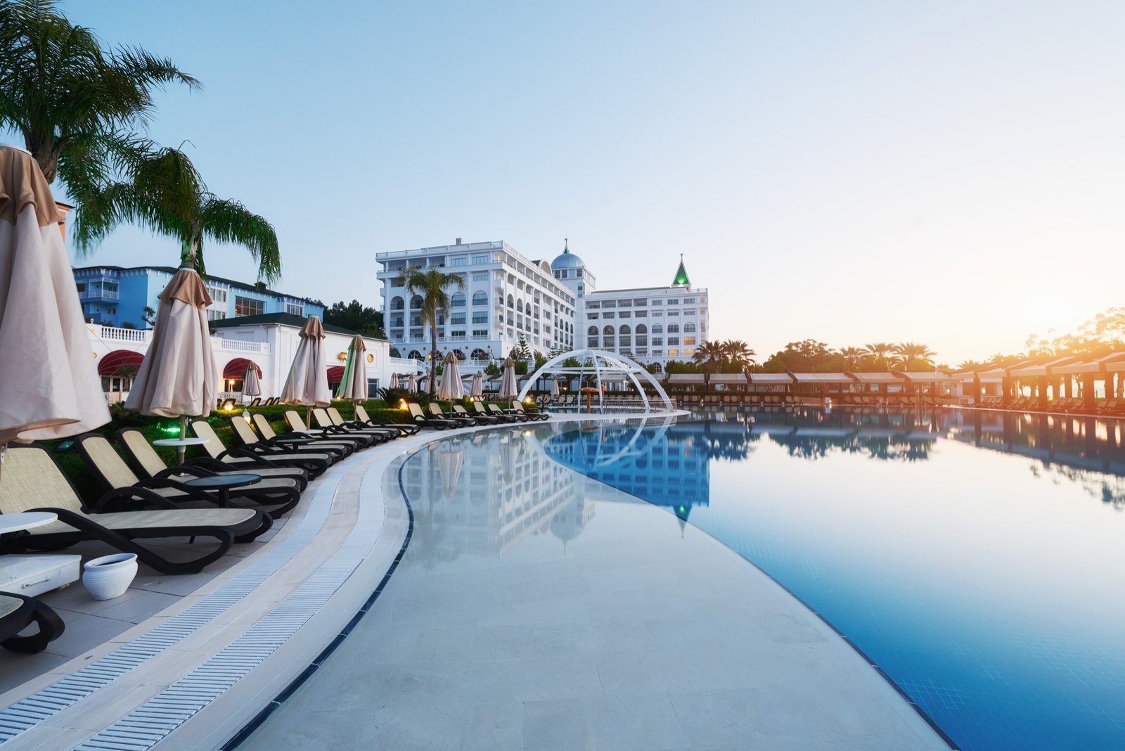 Luxury resort pool with elegant white buildings in the background, surrounded by palm trees and sunbeds.