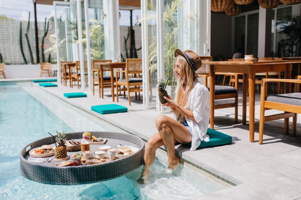 A woman enjoying a tropical breakfast by the poolside, sipping from a pineapple while relaxing with her feet in the water.