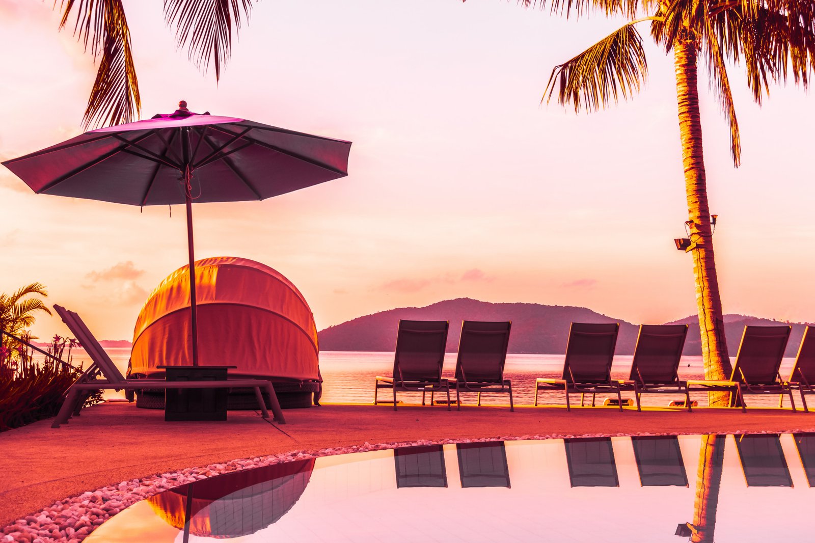 Romantic sunset by the pool, with cozy lounge chairs and palm trees reflecting on the water’s surface.