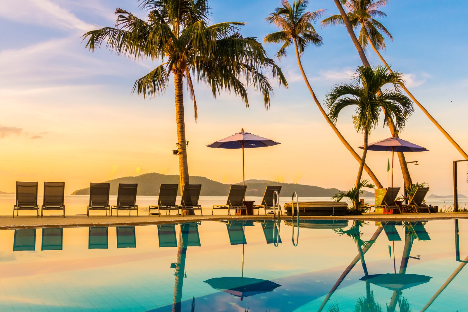 A stunning infinity pool surrounded by palm trees at sunset, with lounge chairs and umbrellas overlooking the ocean.